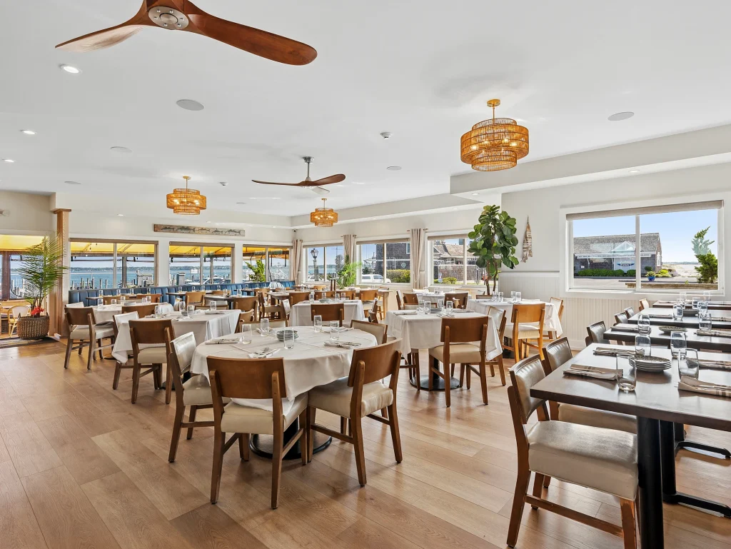 A modern dining room with wooden tables and chairs set with white tablecloths, against large windows with views of a waterfront.