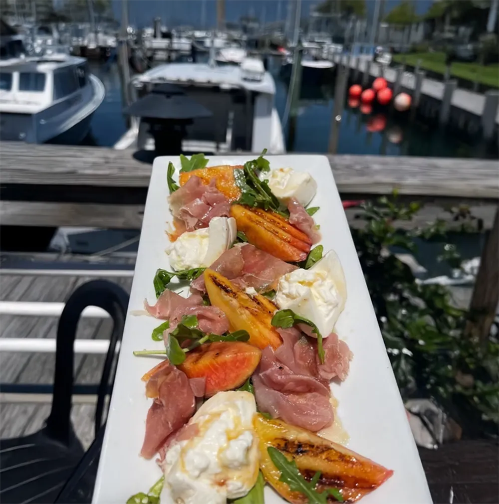 A platter of salad with prosciutto, tomatoes, mozzarella, and arugula, with a marina in the background.