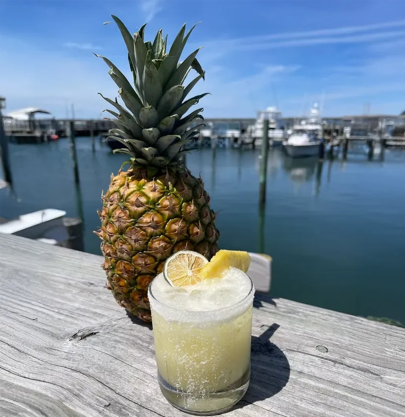 A pineapple and a glass of yellow beverage on a wooden dock by a marina.
