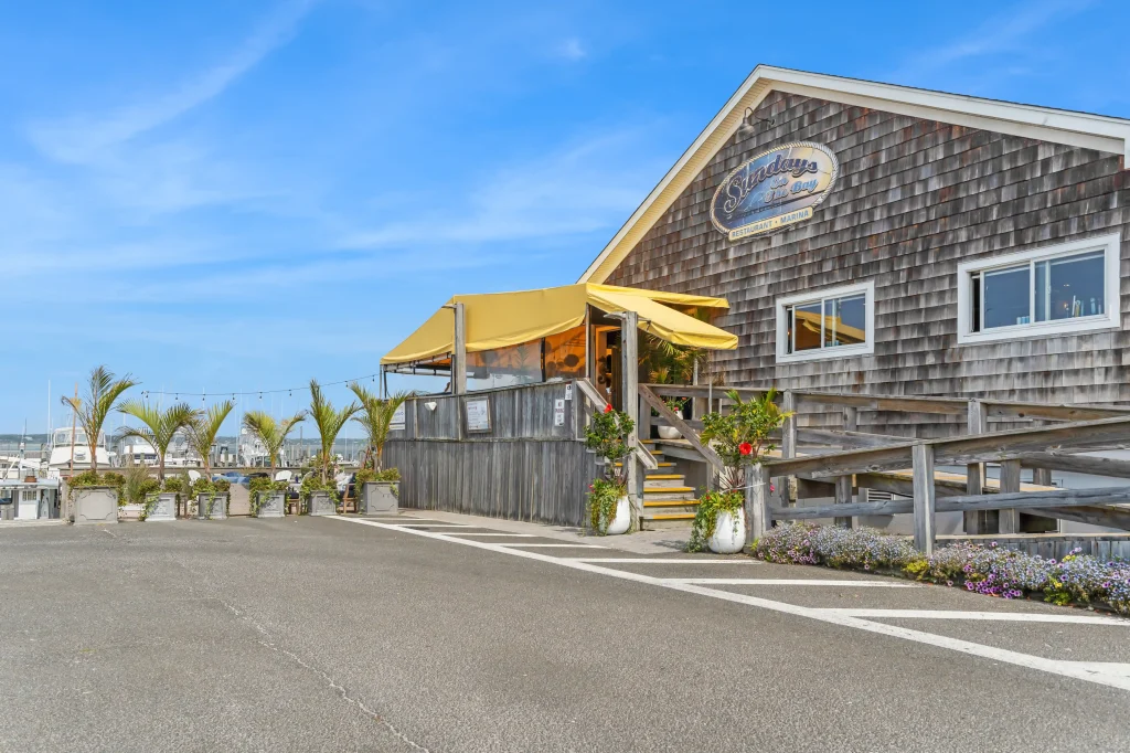 A shingled building with a yellow awning and vibrant plants, next to a marina with boats under a blue sky.