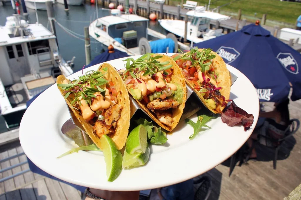 Three fish tacos with grilled fish, vegetables, sauce, and garnishes on a plate by a marina.