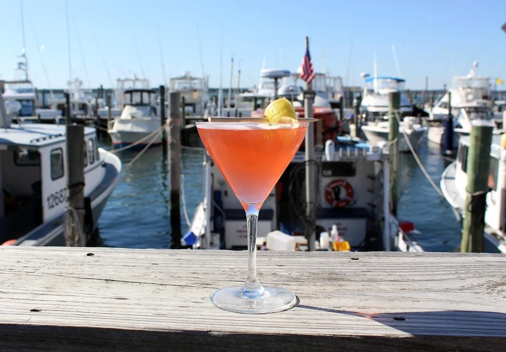A martini glass with an orange-pink cocktail and lemon twist, set in a marina backdrop.
