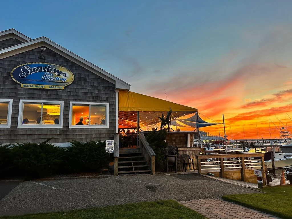 Waterfront restaurant with a sign "Sundays On The Bay" beside a marina at sunset.
