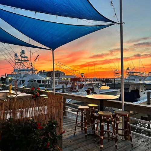 A marina at sunset with boats, a wooden deck with stools, blue shade sails, and vibrant sky.
