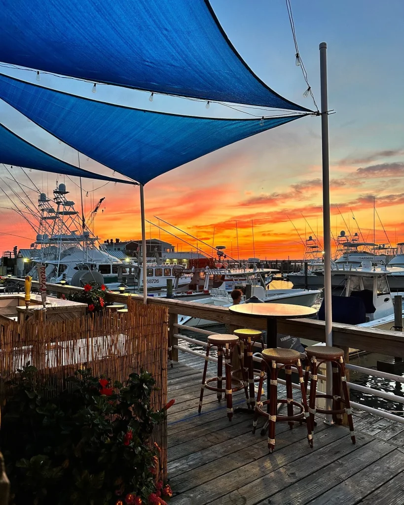A marina at sunset with boats, a wooden deck with stools, blue shade sails, and vibrant sky.