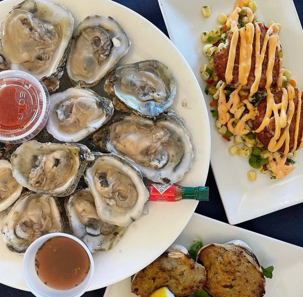 A platter of raw oysters with cocktail sauce and hot sauce, crab cakes with sauce on a bed of vegetables, and baked stuffed clams with a lemon wedge.