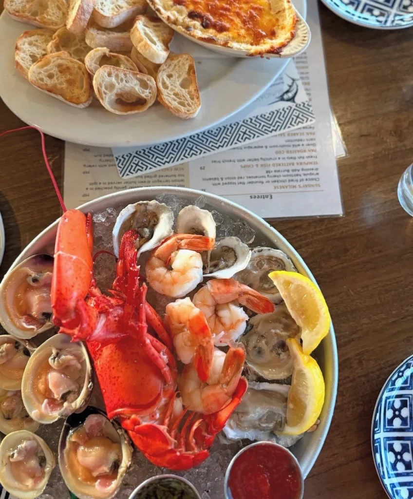 Seafood platter with lobster, shrimp, clams, oysters, and lemons, served next to a plate of bread and a baked dish.
