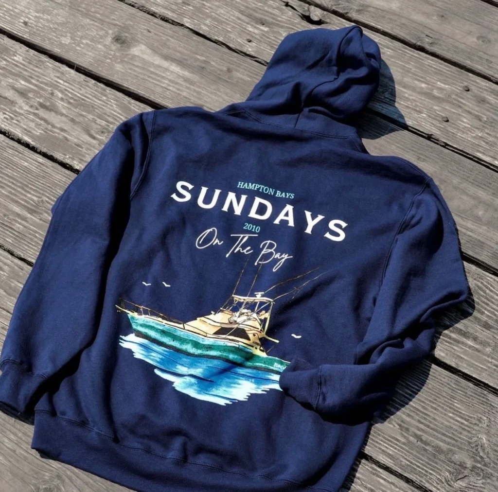 Navy blue hoodie with a boat graphic and text laid on a wooden deck.